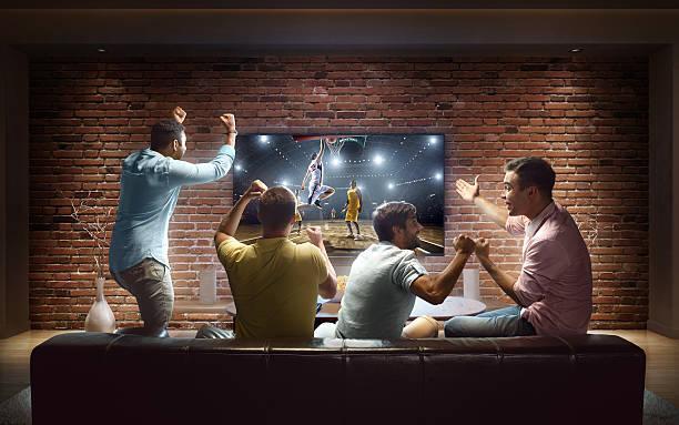 Students watching Basketball game at home :biggrin:A group of young male friends are cheering while watching Basketball game at home. They are sitting on a sofa in the modern living room faced to a big TV set on the front wall. basketball game stock pictures, royalty-free photos & images