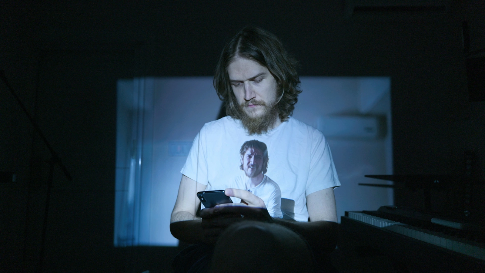 Bo Burnham looks at his phone as a video of himself is projected onto his t-shirt and the wall behind him.