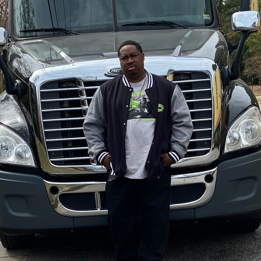 Trucking company owner Larry Gilliam in black and gray jacket standing in front of his 2015 Freightliner Cascadia.