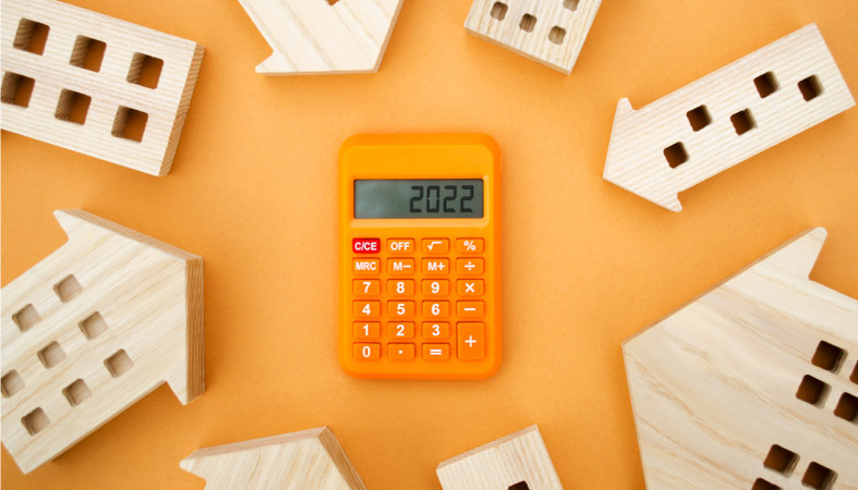 An orange calculator displaying “2022,” against an orange background, surrounded by little wooden house figurines that also double as pointing arrows — representing the concept of trying to figure out the upcoming 2022 housing market rates.