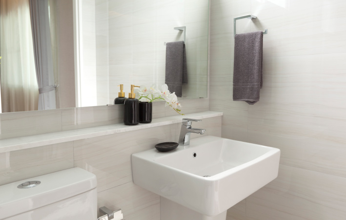 A half bathroom with the white toilet and pedestal sink on the same wall. Above them is a wall-length mirror and shelf with a soap set. On the adjacent wall is a gray hanging hand towel.