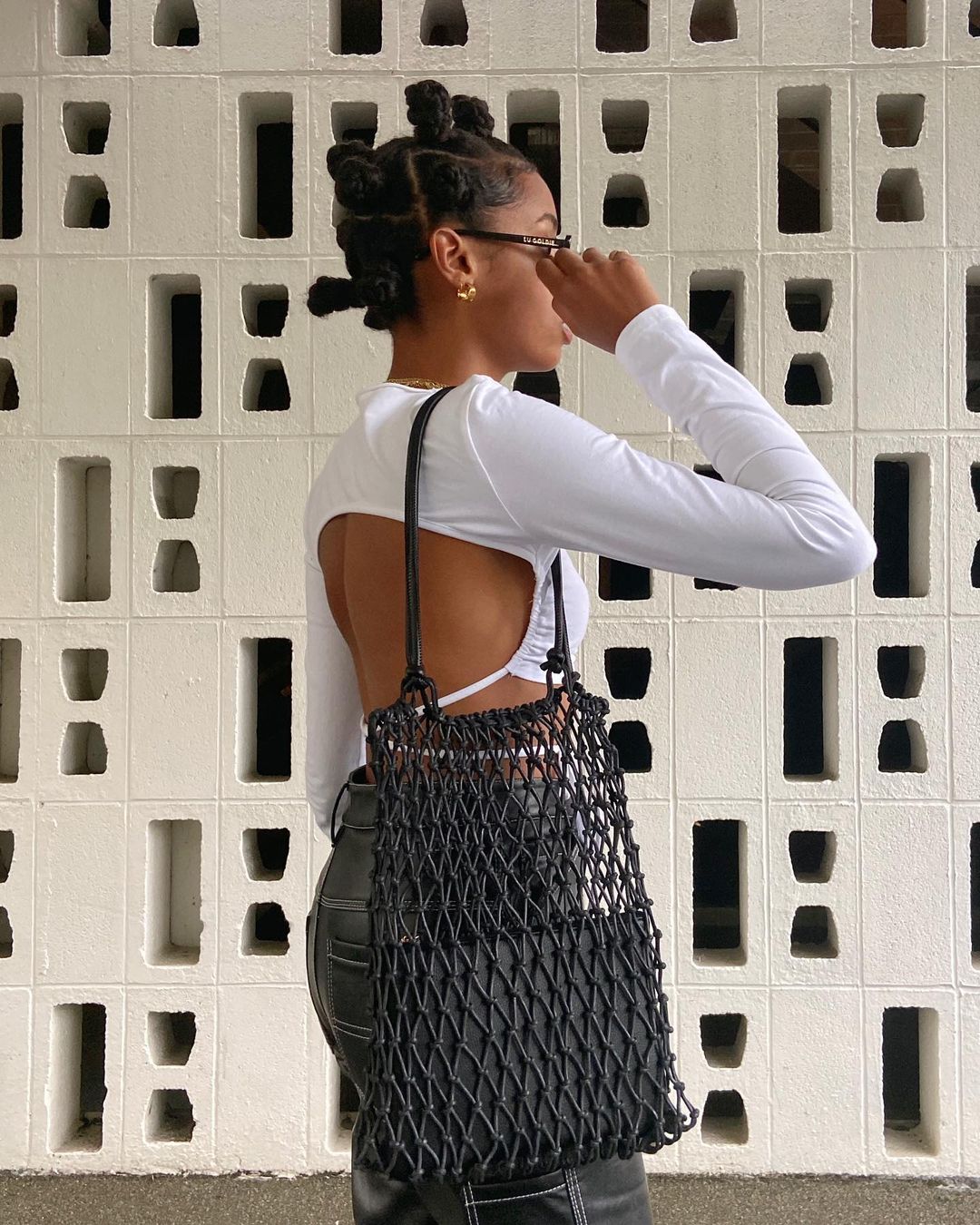 Mulher negra, de cabelos pretos e presos. Ela usa um cropped de frio branco, com calça de couro. Sua bolsa é uma net bag, toda aberta em rede.