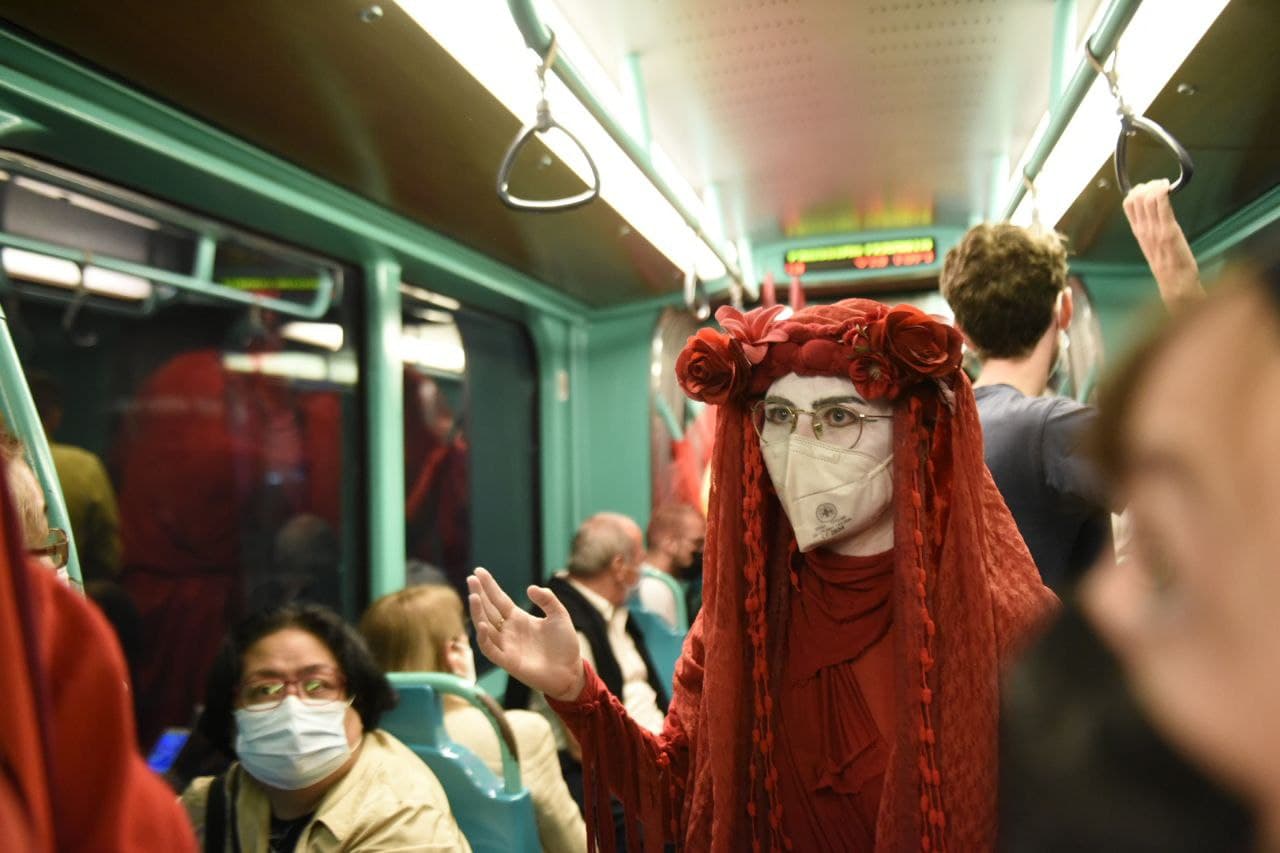A red rebel in white facepaint and red robes stands in a train carriage full of commuters.