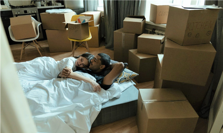 A couple is sleeping on a mattress on the floor of their new apartment. Moving boxes are stacked all around them.