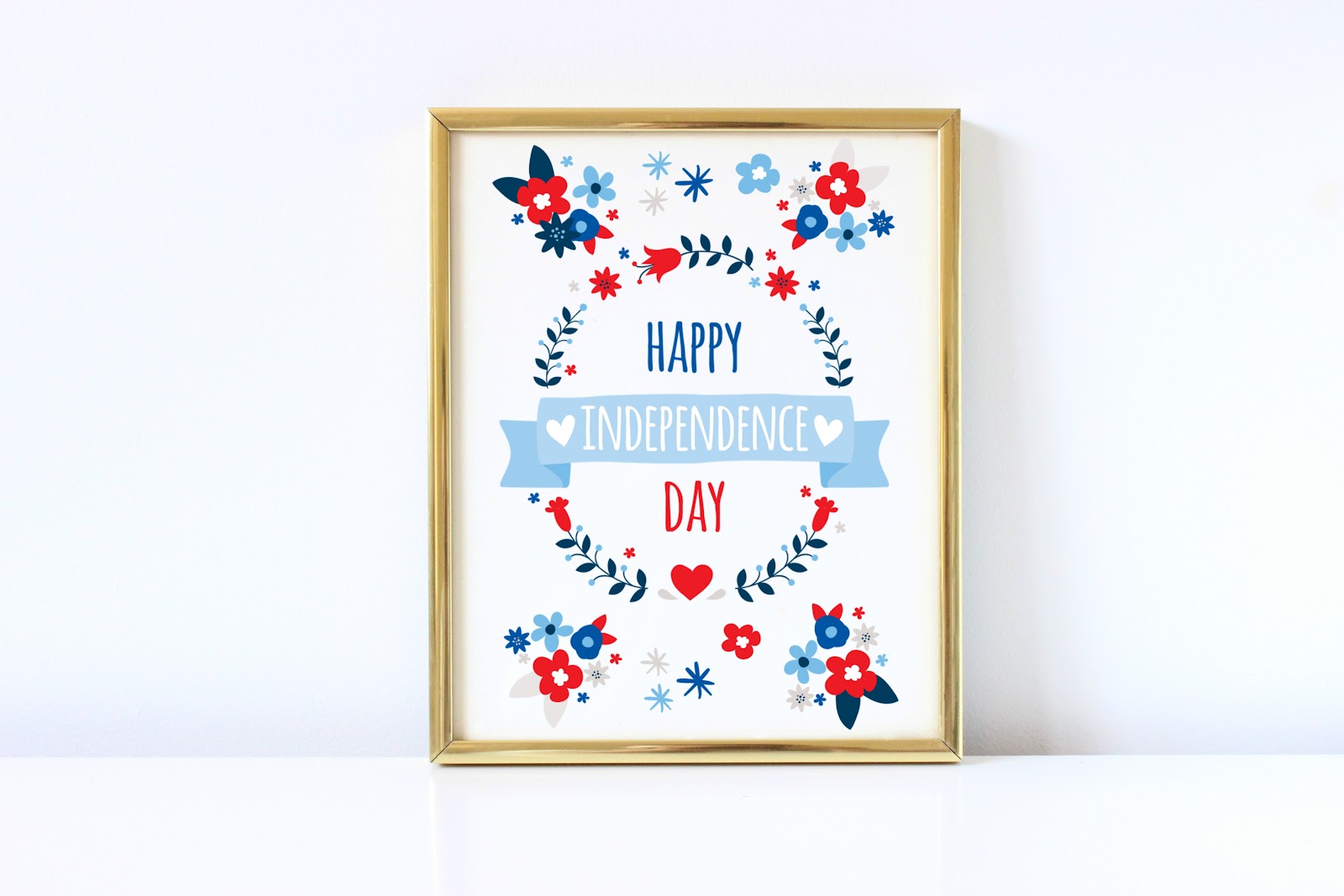 A Fourth of July wall art piece is displayed in a gold frame against a white wall. The print has red, white, and blue flowers and text in the center that reads, “Happy Independence Day.”