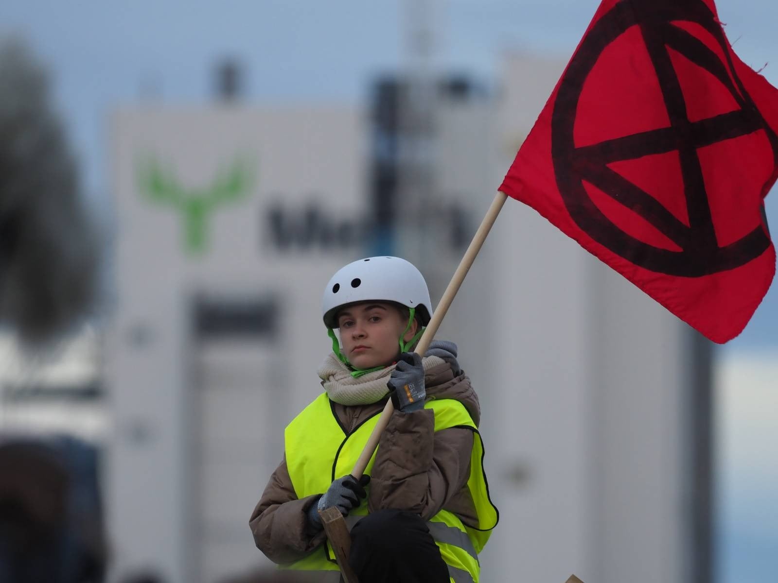 A young rebel holds an XR flag atop a tripod outside a pulp mill.