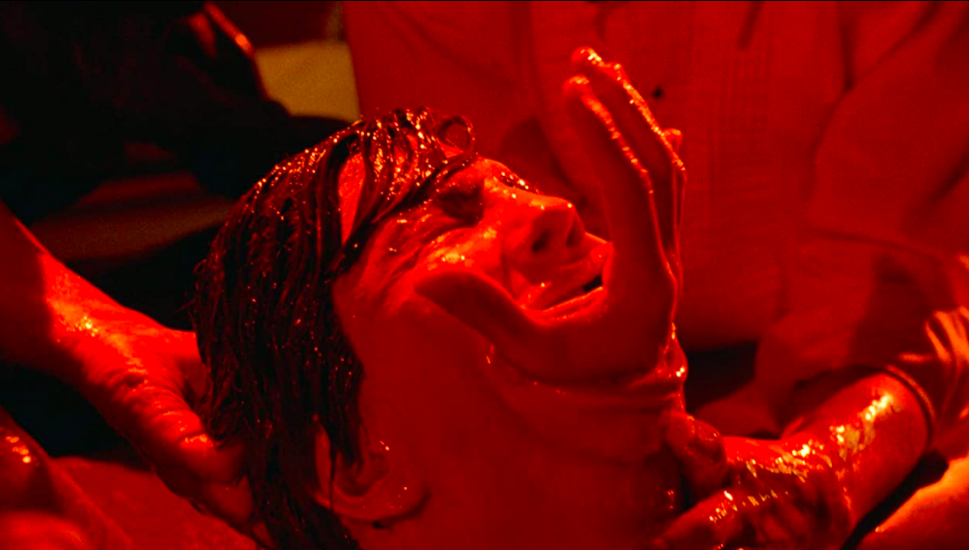 Society (1989). A grotesque body horror close-up of a man's face, bathed in bright red lights and soaked with blood. A human hand is protruding from his mouth as if reaching out from his throat, the man's face is contorted with pain.