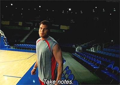 A man standing on the edge of an indoor sports court. He leans in close to the camera, making eye contact, and speaks to it. The caption reads "Take notes."