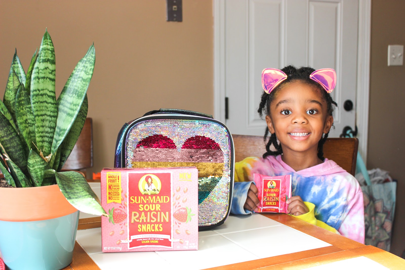 Girl with colorful shirt holding Sun-Maid Sour Raisin Snacks at table with lunchbox 