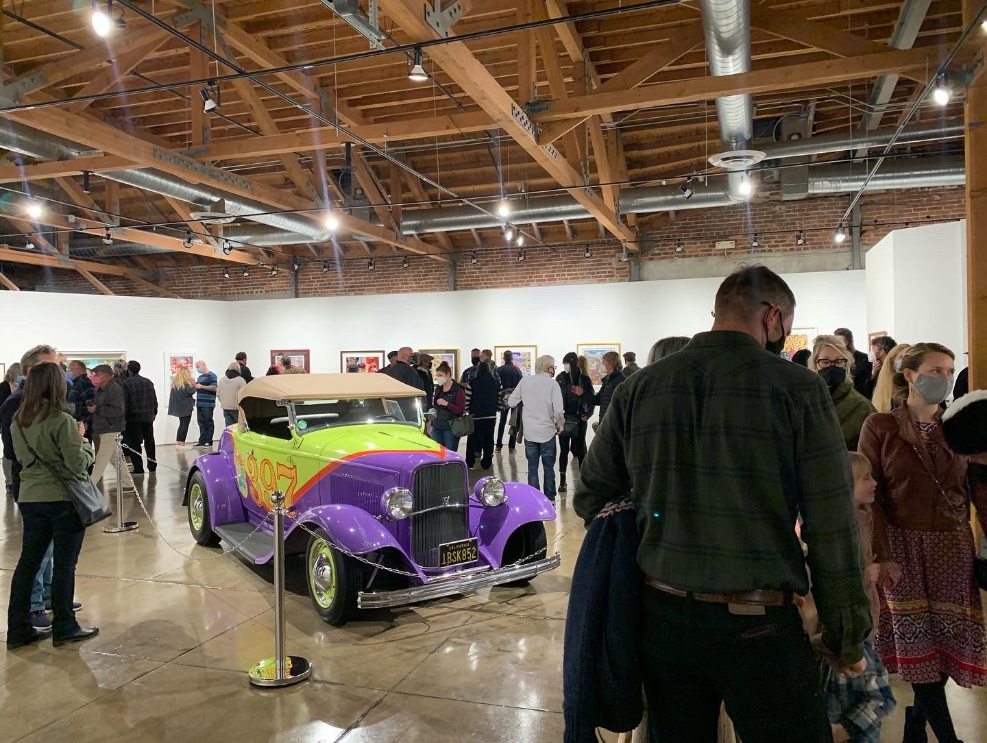 A picture of a group of people observing the artworks on the walk and a car with the colors yellow and purple in the center of the room