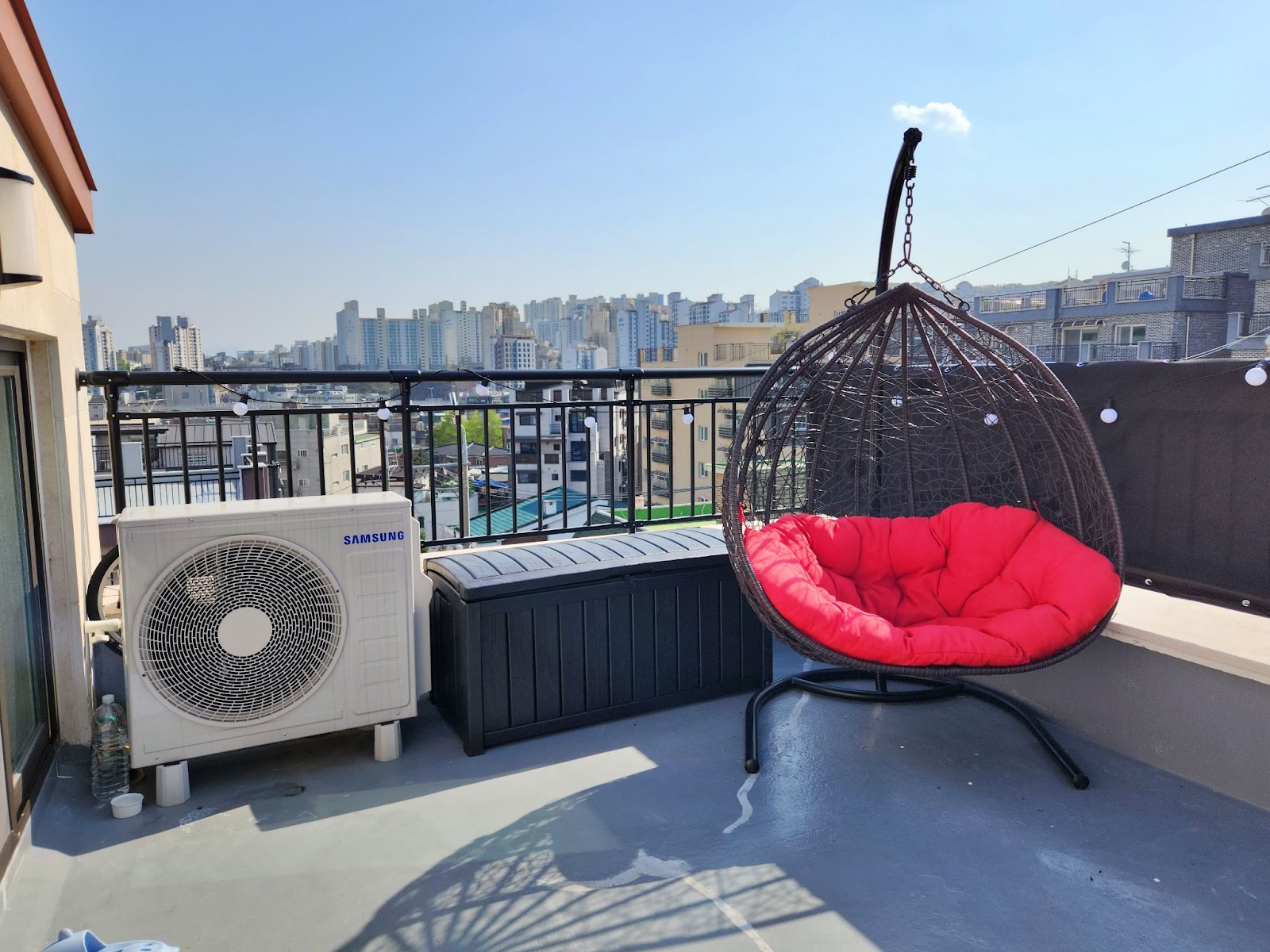My rooftop area with a handing bamboo chair with red cushions, with the Seoul cityscape in the background.