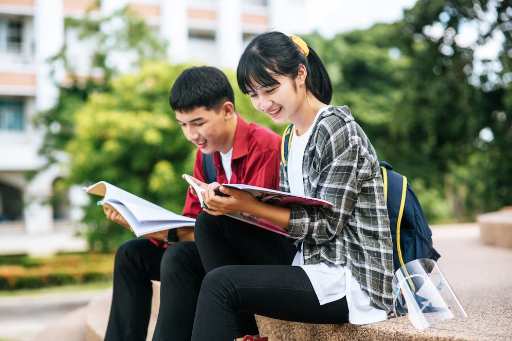 A couple of students sitting on a bench reading books

Description automatically generated