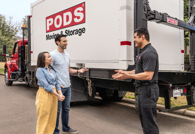 A PODS driver is explaining the process of relocating the moving and storage container to a couple. They are all standing in the street with a loaded container on a PODS truck behind them.