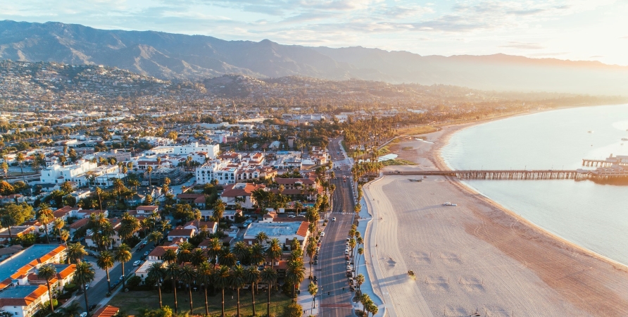 An early morning aerial view of the city of Santa Barbara, its beachfront, and pier, with the mountains in the background.
