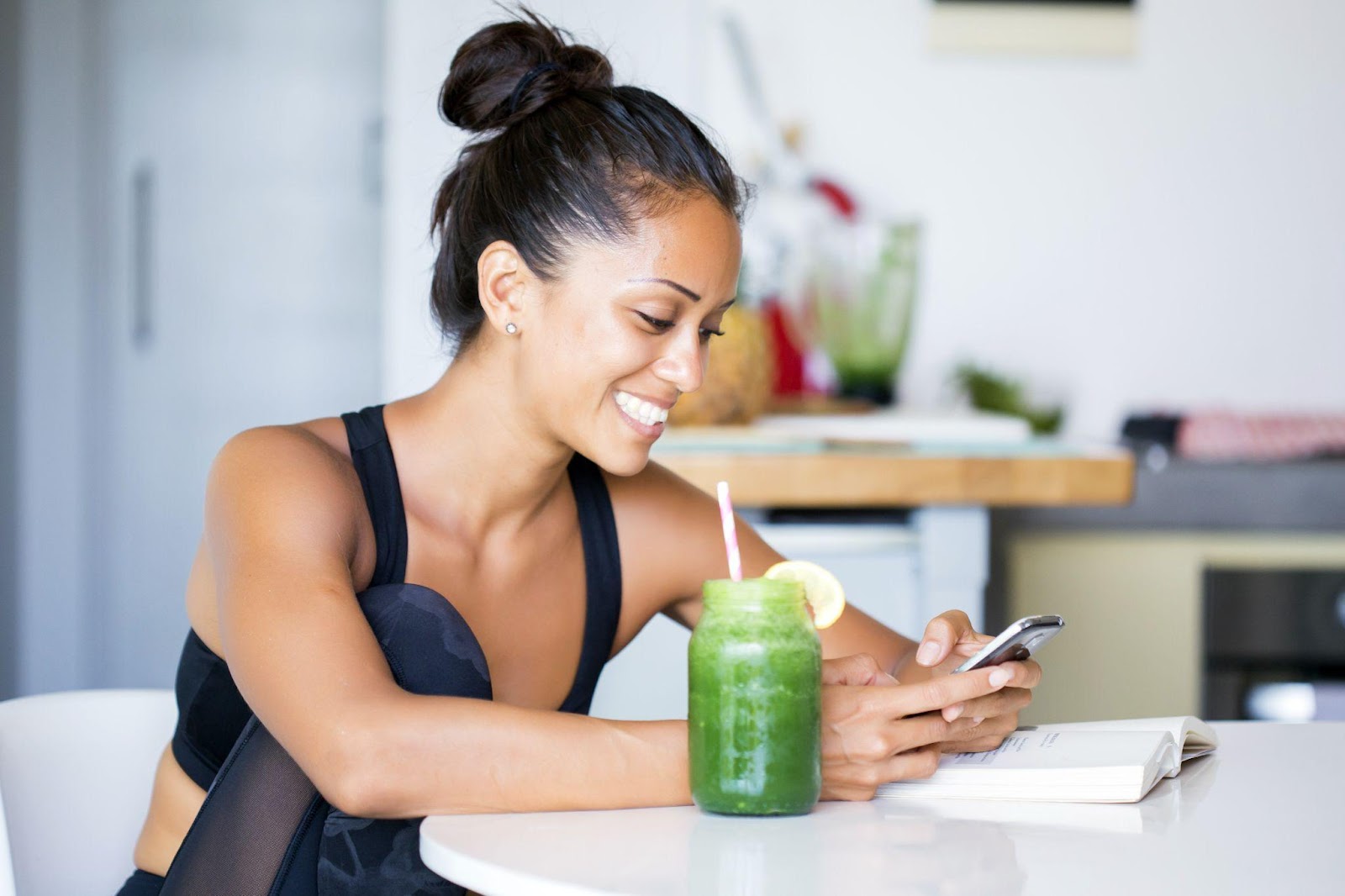 Mujer bebiendo un zumo verde hecho en casa, con ropa deportiva, escribiendo en su teléfono mientras está sentada en la mesa de su cocina.