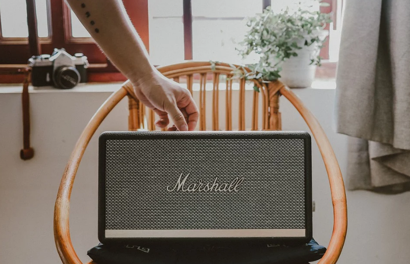 A person’s hand adjusting a knob on a Marshall amplifier which is placed on a wooden chair. In the background, there is a camera on a windowsill and light entering through the window, creating a warm ambiance.