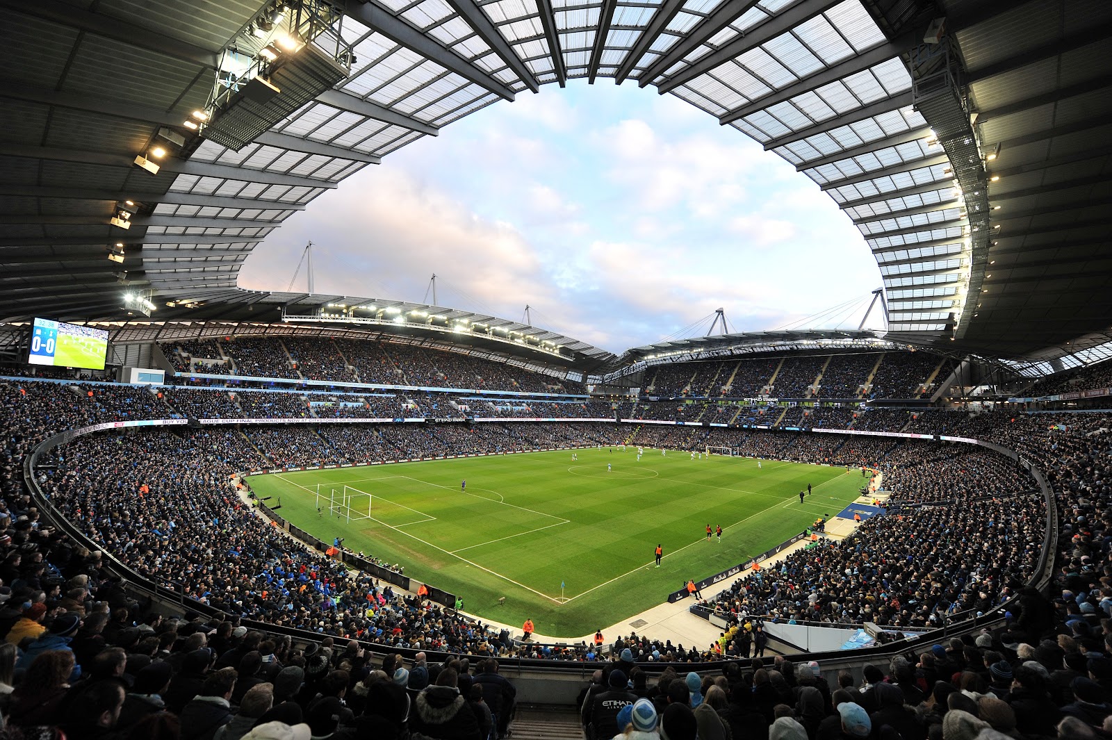 manchester etihad stadium busy during a large football game. footballers playing on the pitch, sunset, the stadium is illuminated. wide angle shot from the upper rows at the stadium.