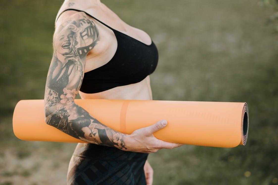 Side view of crop anonymous tattooed female in sports clothes with rolled yoga mat standing on lawn