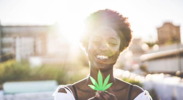 Young african girl holding marijuana leaf - Cannabis medicine, real people, healthy lifestyle and ecology concept - Focus on her face  smoking CBD happy stock pictures, royalty-free photos & images