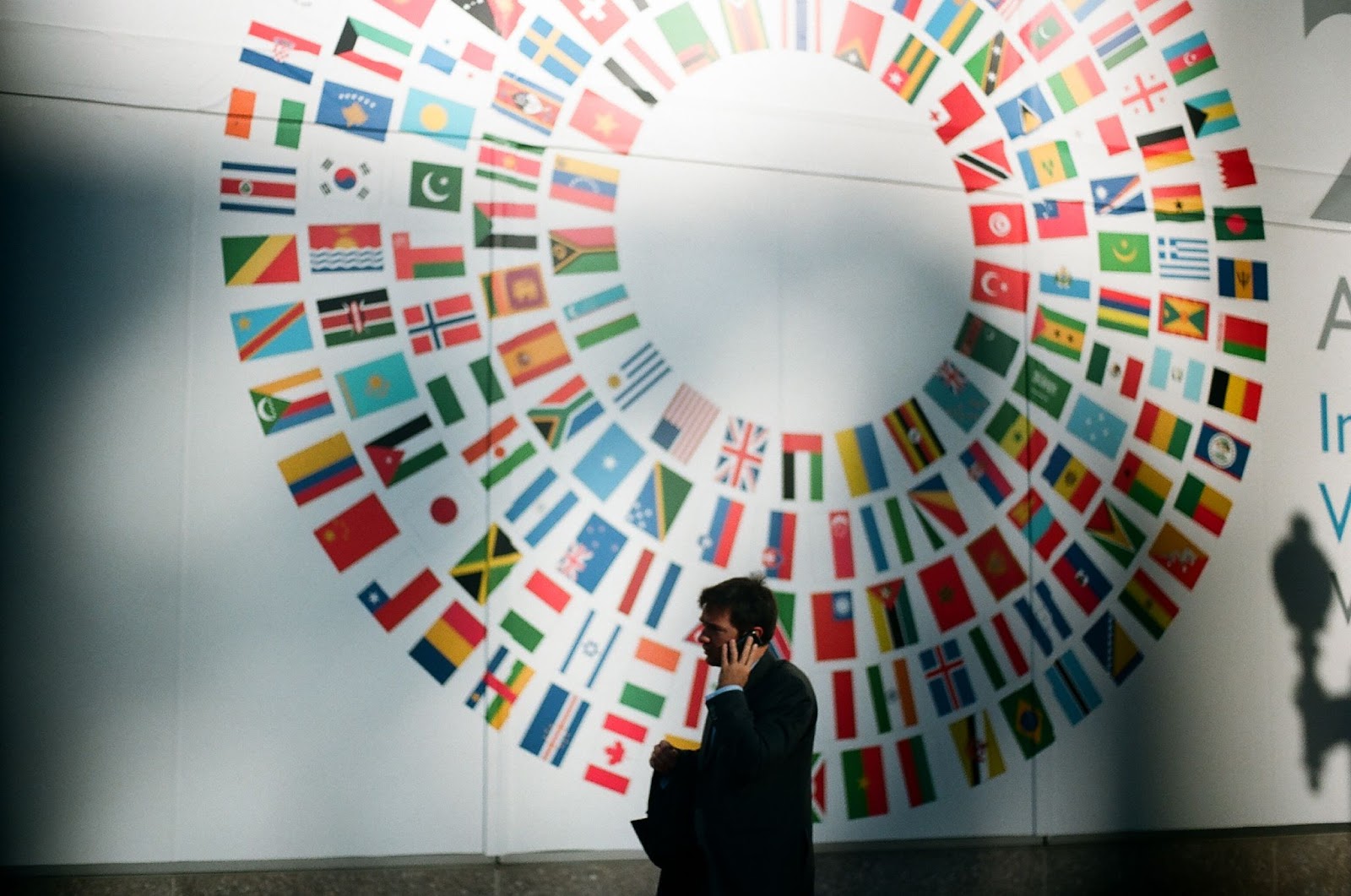 Homem branco de terno preto caminhando em frente a parede branca com bandeiras de diversos países pintadas em círculo