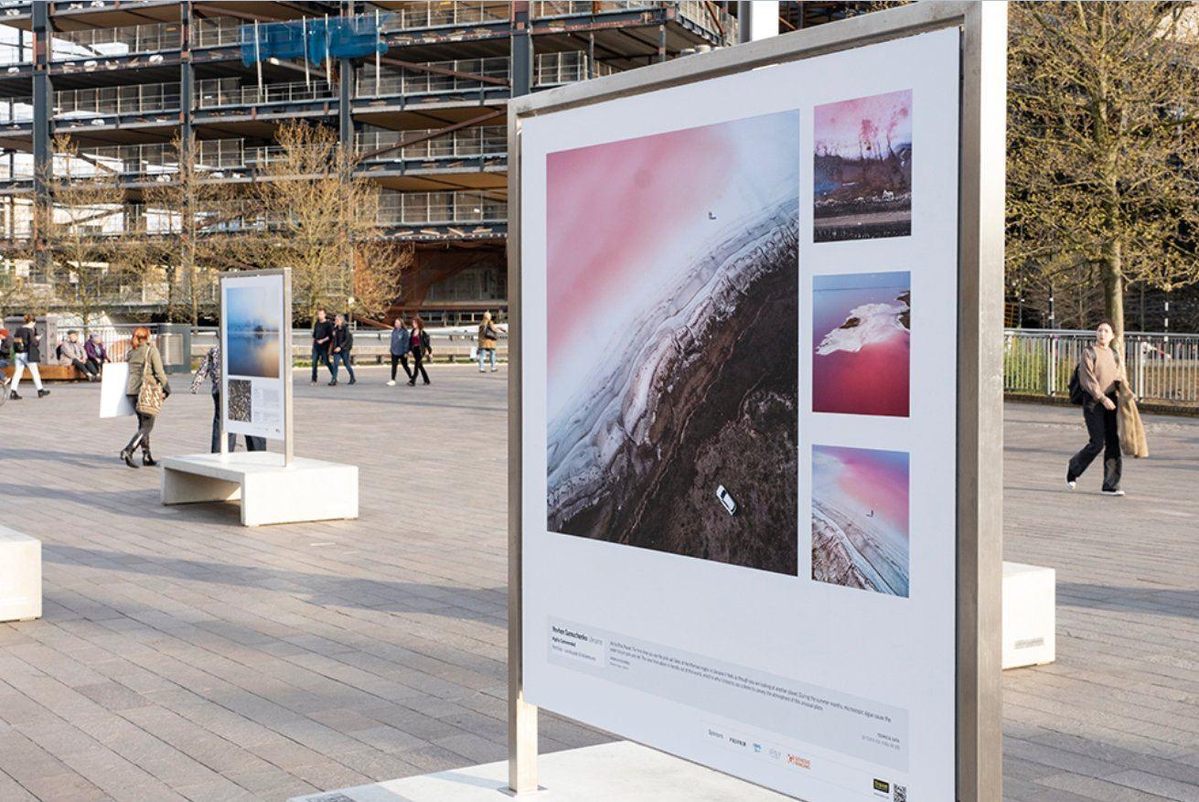 An image of an outdoor exhibition featuring some of Yevhen's work. Concrete benches with his images printed on boards are dotted throughout a busy plaza.