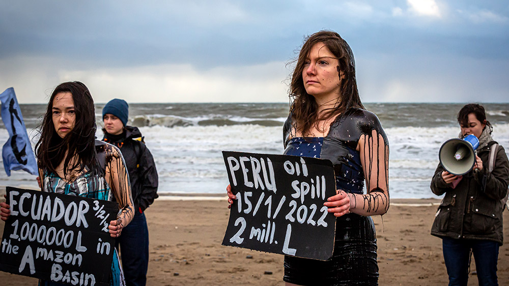 Two young women covered in fake oil stand on a beach holding signs stating country names and the size of the oil spill there.
