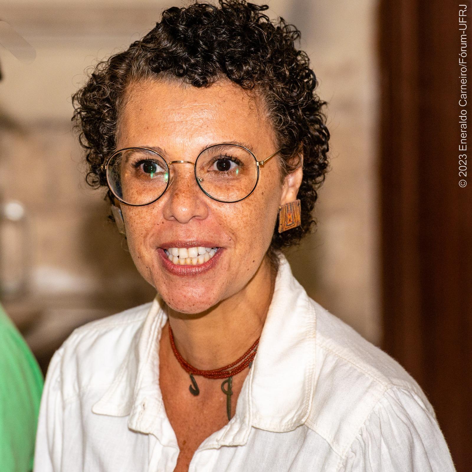 Na foto, Marcia Cabral aparece usando óculos de grau redondos, blusa branca, colar vermelho e brinco no formato quadrado.