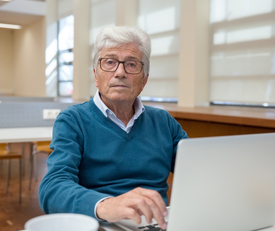 A imagem mostra um idoso de cabelos brancos que olha para a câmera enquanto digita em seu notebook. O senhor usa um óculos preto e um colete de lã azul-turquesa por cima de uma camisa branca.