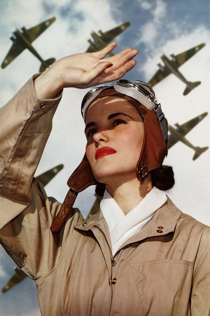 Uma mulher vestida com roupas de aviadora do anos 40 olha para o céu enquanto aviões passam.