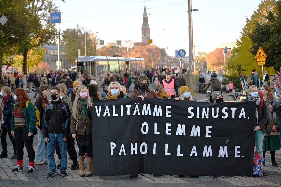 Young rebels stand in a line in a busy square in Helsinki holding up a large black banner.