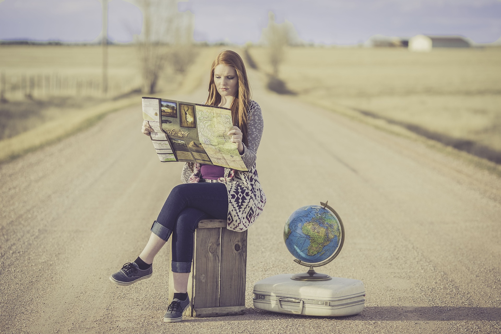 Girl sitting in a road reading a map with a globe and suitcase next to her