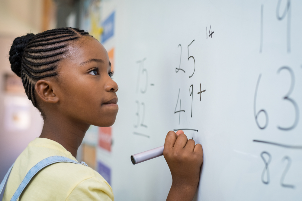 Menina negra do ensino fundamental está fazendo uma conta de matemática em uma lousa branca.