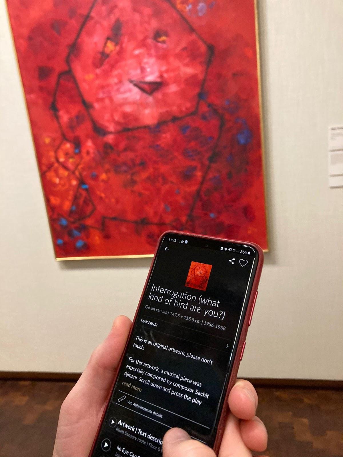 A photo of the artwork Interrogation (What Kind of Bird Are You?) (1956-1958) by Max Ernst and a phone held-in hand in front of it. On the screen of the phone is the digital app of the museum that contains the musical piece composed for the piece by composer Sachit Ajmani that the visitors can listen to.