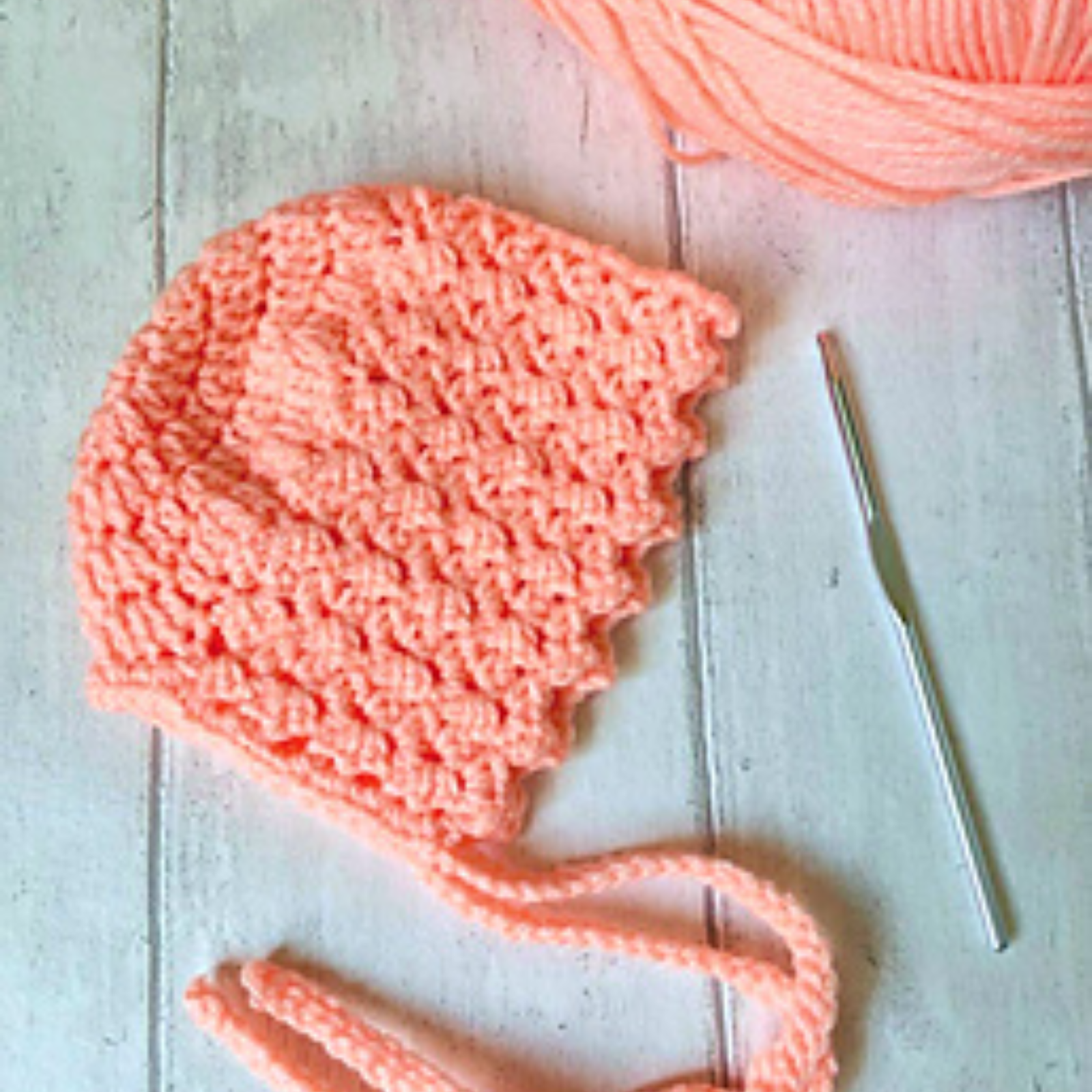 peach colored baby bonnet sitting on wooden background with hook and yarn