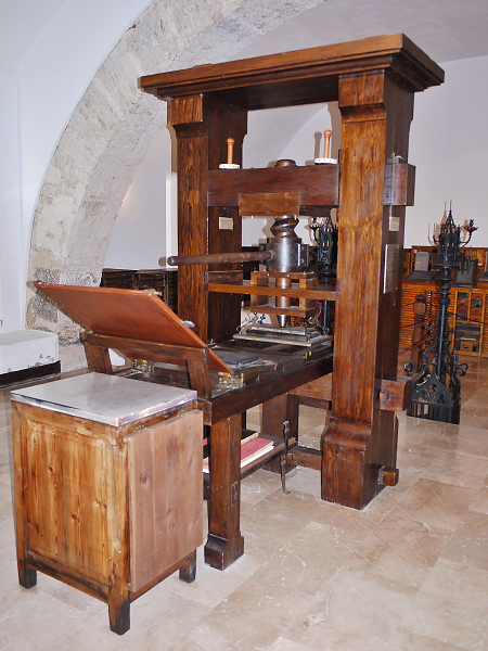 Photograph of a replica of an early printing press. It is made of wood with a significant metal part in the middle, attached to a large lever.
