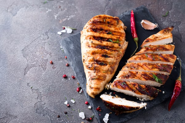 Two grilled chicken breasts, one whole and one sliced, with wild chili and garlic placed on a stone board