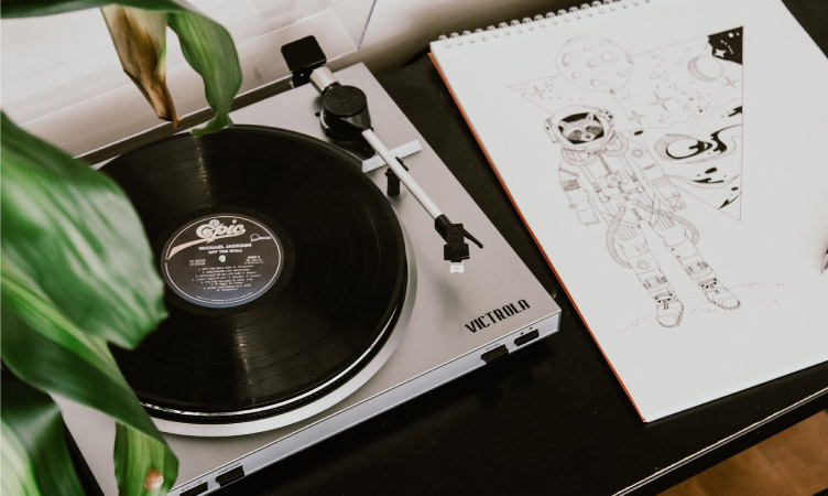 A Victrola record player with a black vinyl record on it. There is a drawing pad beside it with a black and white drawing of a racoon in a spacesuit and a geometric shape with images of outer space drawn inside it. A houseplant’s leaves are reaching over the record player.