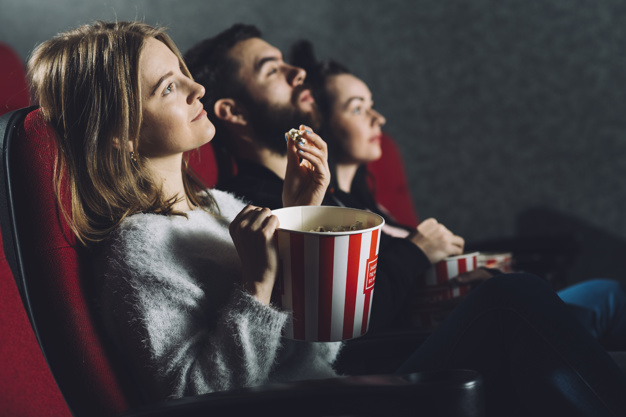 Três pessoas sentadas em poltronas de cinema assistindo um dos melhores tipos de filmes comendo pipoca. 