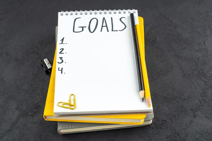 A bottom view of a notepad with 'list of goals' written on it, accompanied by a pencil sharpener, black and yellow pencils, gems, and clips, on a dark background.