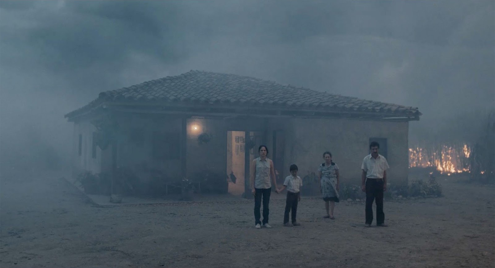 A still from 'Land and Shade' (2015), a family standing outside a house enflamed in smoke. 