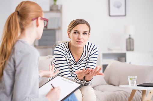 A white woman sitting in a therapy couch leaning forward towards the therapist who has a notebook and taking notes. This photo represents seeking counseling for mental health needs such as anxiety, depression, OCD, relationship issues. Anxiety treatment in Los Angeles, CA can help with coping skills by talking to an anxiety therapist. | 93020 | 94513 91356 | 91020