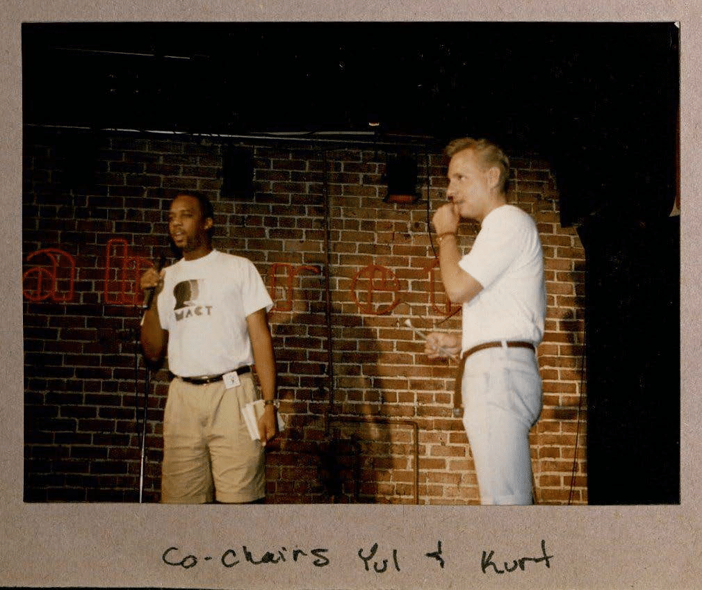 Yul Stell and Kurt M., co-chairs of Men of All Colors Together-KC from 1992–1993, pictured above ([Scrapbook clippings from BWMT/MACT-KC], ca. 1992-1993). Both men stand in front of a brick wall, wearing the MACT-KC t-shirt. Yul speaks into a microphone as Kurt looks onward.