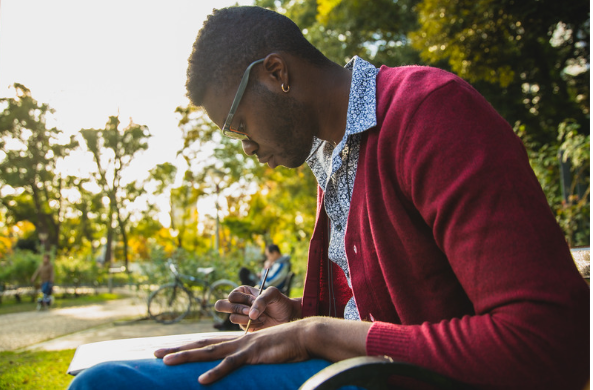 Photo shows a student studying (writing on a notebook) at a park.