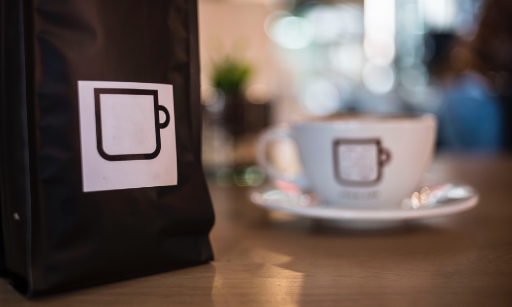 Black multilayer kraft paper coffee bag with white label sitting on counter with white ceramic coffee cup and saucer in background. 