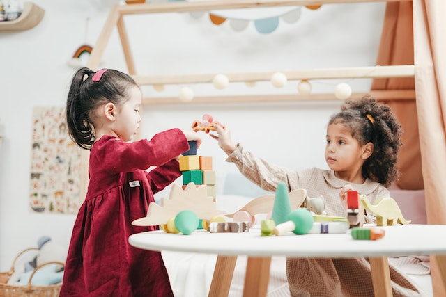 young girl in red dress plays with wooden blocks at a table as another girl in a brown dress sits next to her playing with dinosaur toys