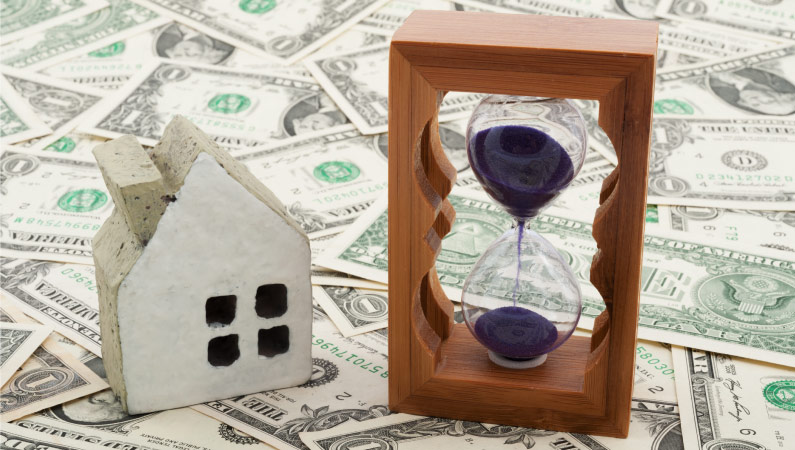 A little figurine of a house beside a wooden and glass hourglass, atop scattered dollar bills — representing the quandary of when to buy or sell your house.
