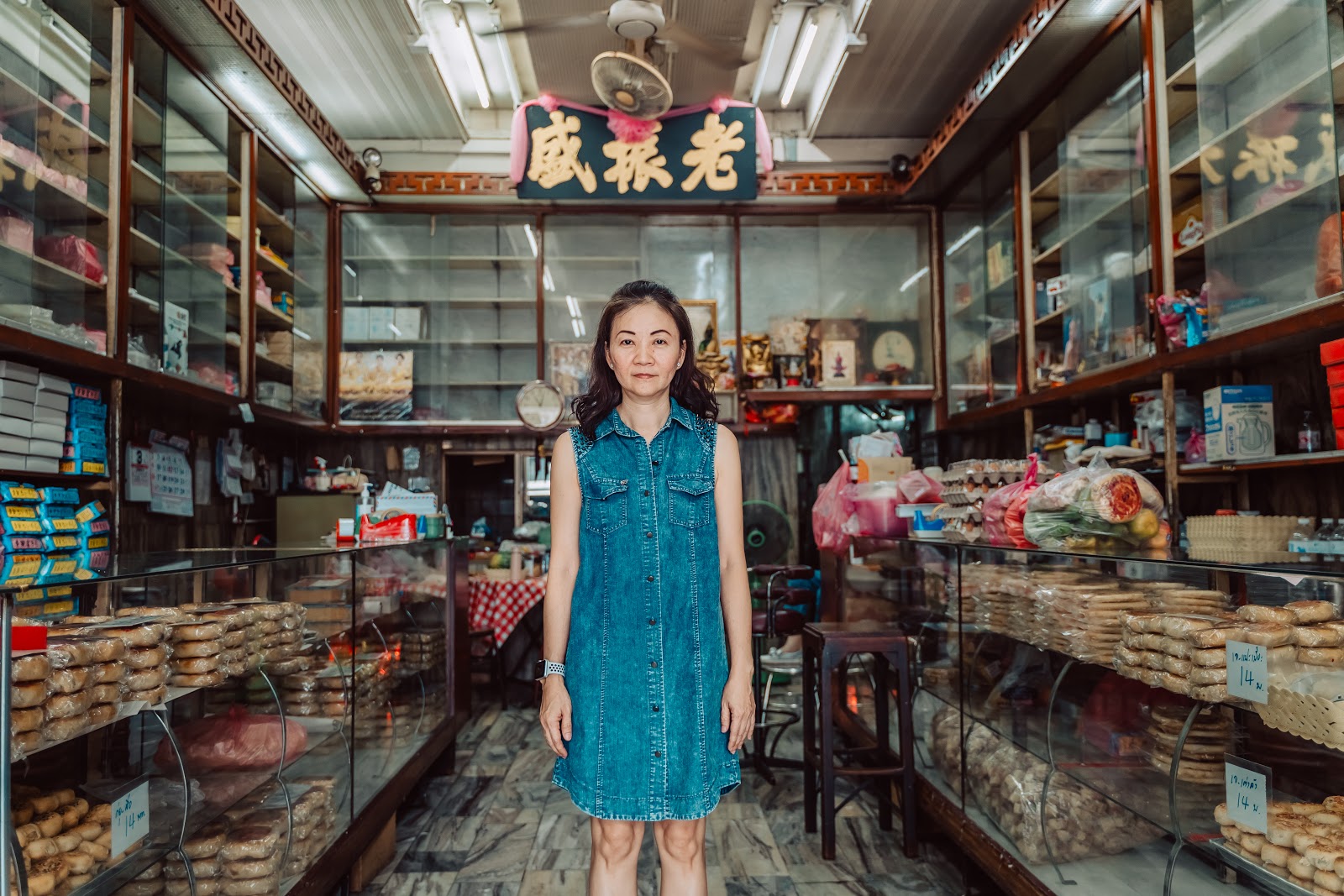 Image 1: 一位穿著牛仔洋裝的女性站在傳統店鋪內，周圍擺滿了各式各樣的商品，店鋪上方掛著寫有「老聚威」的招牌