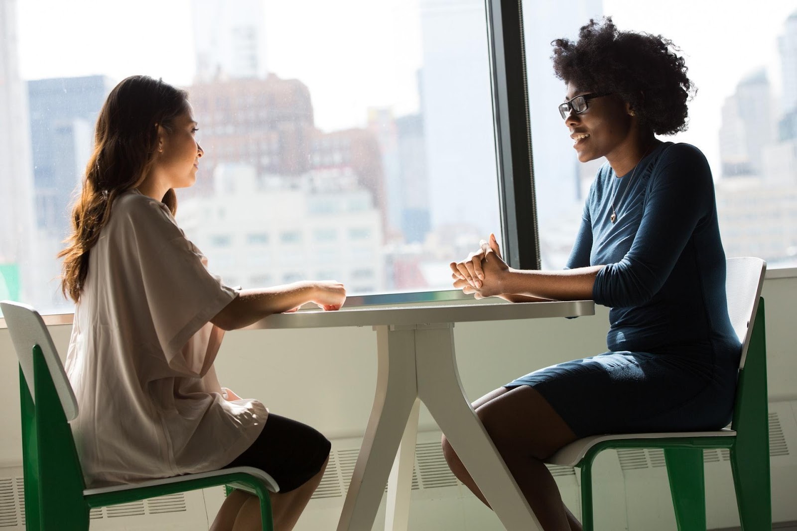 La imagen muestra a dos mujeres sentadas frente a una mesa de madera blanca. Están una frente a otra y se ven desde un ángulo lateral. A la izquierda, hay una mujer blanca con pelo castaño ondulado. A la derecha, hay una mujer negra, con cabello rizado y negro. Ambas están usando ropa más social.