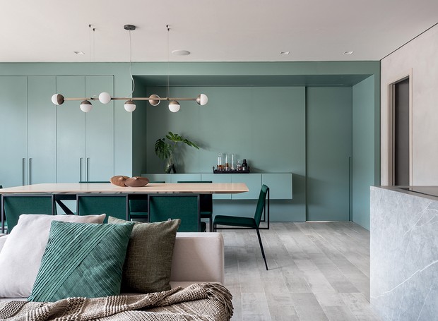 Sala de jantar integrada a sala de estar com armário e aparador de madeira na cor verde, mesa de jantar de madeira com oito cadeiras verdes, sofá branco com almofadas coloridas e piso de madeira