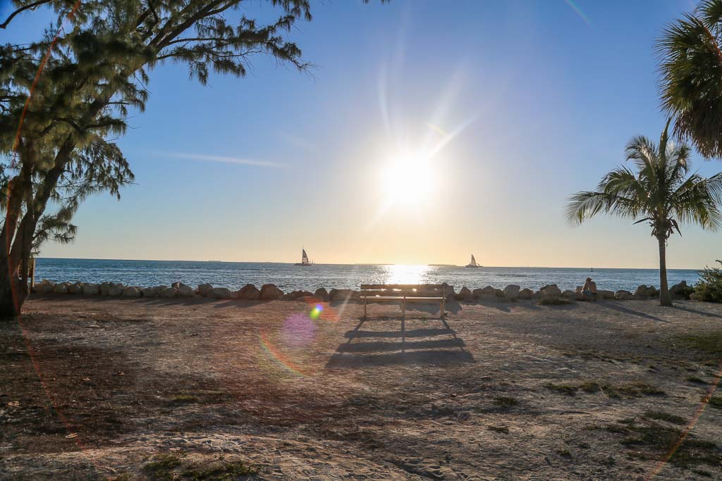 The beginning of sunset from the beach at Zachary Taylor. A sun flare takes up much of the image and trees line the side. There is a sandy beach with a bench and long bench shadow. There is a row of rocks across the photo between the sand and water. Two boats are far-off in the water.